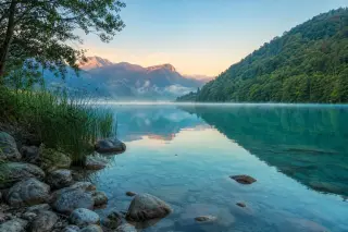 Mist rising over a quiet, stony lakeshore framed by alpine vegetation, turquoise water, and reflected hills near Annecy.