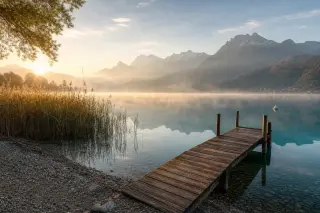 Quiet lakeshore of Annecy at dawn, empty beach with distant mountains reflected in calm water, soft mist and gentle morning light.