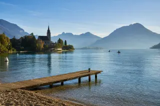 Pebble shoreline at Duingt on Lake Annecy, mountains and a wooden pier, gentle morning light, subtle kayaks in distance, authentic alpine scenery.
