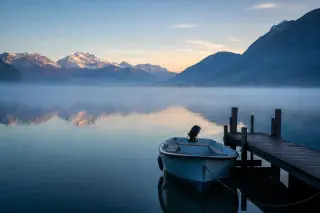 Misty dawn over Lake Annecy with a small boat near a wooden dock, surrounded by Alpine peaks and calm, reflective water.
