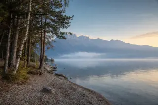 Peaceful lakeshore at Lake Annecy under mist and golden morning light, with distant Alpine mountains and calm water.