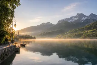 Serene Lake Annecy shoreline at dawn with gentle mist and Aravis mountains in the background, tranquil natural scenery.