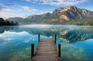 Peaceful Lake Annecy with mountain backdrop and mist, a wooden jetty extending into the water.